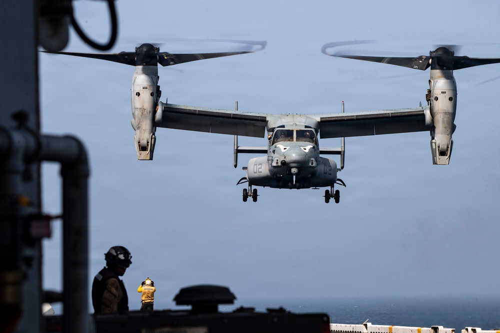 A V-22 Osprey tiltrotor aircraft marked '02' is hovering during an approach to land on what appears to be a naval vessel's flight deck. The rotors are in vertical/helicopter mode, indicating a landing or hover operation. A flight deck crew member in a yellow jersey is visible on deck, appearing to guide the aircraft.