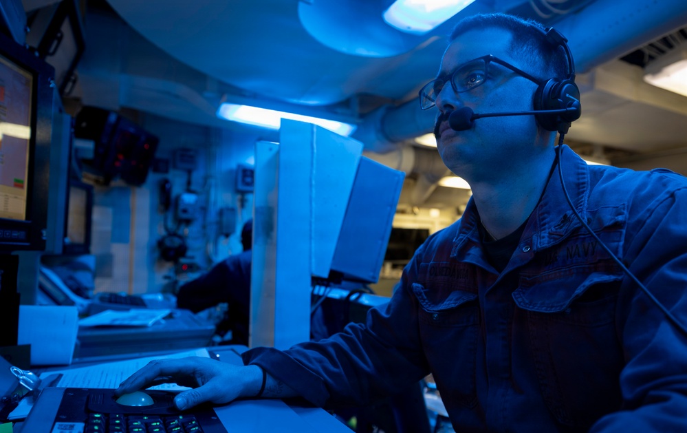 A military personnel member, likely U.S. Navy based on the blue coverall uniform, is seated at a control console in what appears to be a combat information center (CIC) or operations room aboard a naval vessel. The space is illuminated by blue lighting, which is characteristic of low-light operational environments on warships. The sailor is wearing a communications headset and glasses while monitoring screens and operating control equipment.