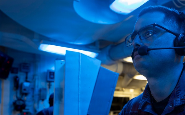 A military personnel member, likely U.S. Navy based on the blue coverall uniform, is seated at a control console in what appears to be a combat information center (CIC) or operations room aboard a naval vessel. The space is illuminated by blue lighting, which is characteristic of low-light operational environments on warships. The sailor is wearing a communications headset and glasses while monitoring screens and operating control equipment.