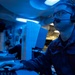 A military personnel member, likely U.S. Navy based on the blue coverall uniform, is seated at a control console in what appears to be a combat information center (CIC) or operations room aboard a naval vessel. The space is illuminated by blue lighting, which is characteristic of low-light operational environments on warships. The sailor is wearing a communications headset and glasses while monitoring screens and operating control equipment.