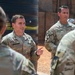 A group of U.S. Army soldiers in OCP (Operational Camouflage Pattern) uniforms are gathered in what appears to be an outdoor forward operating base or military installation. The central figure, identified by name tape as 'CRAWFORD,' is speaking to the group, while another soldier identified as 'CASTRO' stands nearby listening. The setting appears to be in an arid environment based on the red dirt ground and infrastructure visible in the background.