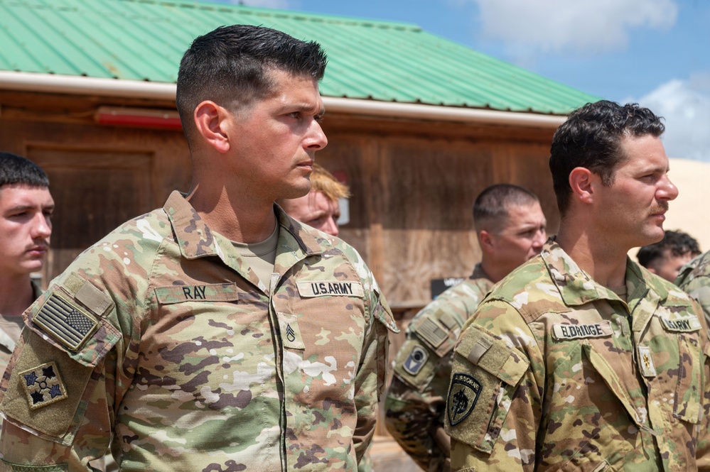 A group of U.S. Army soldiers in Operational Camouflage Pattern (OCP) uniforms are standing in formation, appearing to be at attention during a military ceremony or event. Two soldiers are prominently visible in the foreground, identified by name tapes as 'RAY' and 'ELDRIDGE.' The setting appears to be an outdoor military installation with a building featuring a green metal roof in the background.