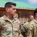 A group of U.S. Army soldiers in Operational Camouflage Pattern (OCP) uniforms are standing in formation, appearing to be at attention during a military ceremony or event. Two soldiers are prominently visible in the foreground, identified by name tapes as 'RAY' and 'ELDRIDGE.' The setting appears to be an outdoor military installation with a building featuring a green metal roof in the background.