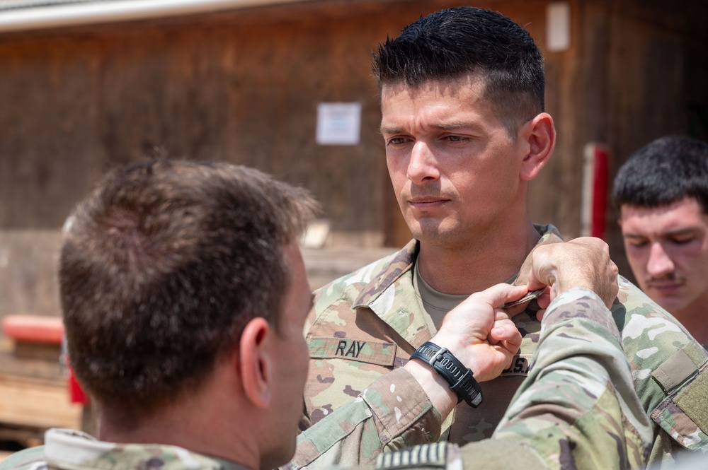 A military ceremony appears to be taking place outdoors, where one soldier is pinning something onto the uniform collar of another soldier identified by the name tape 'RAY'. The recipient soldier is standing at attention while a third soldier observes in the background. The setting appears to be a forward operating base or military installation.