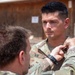 A military ceremony appears to be taking place outdoors, where one soldier is pinning something onto the uniform collar of another soldier identified by the name tape 'RAY'. The recipient soldier is standing at attention while a third soldier observes in the background. The setting appears to be a forward operating base or military installation.