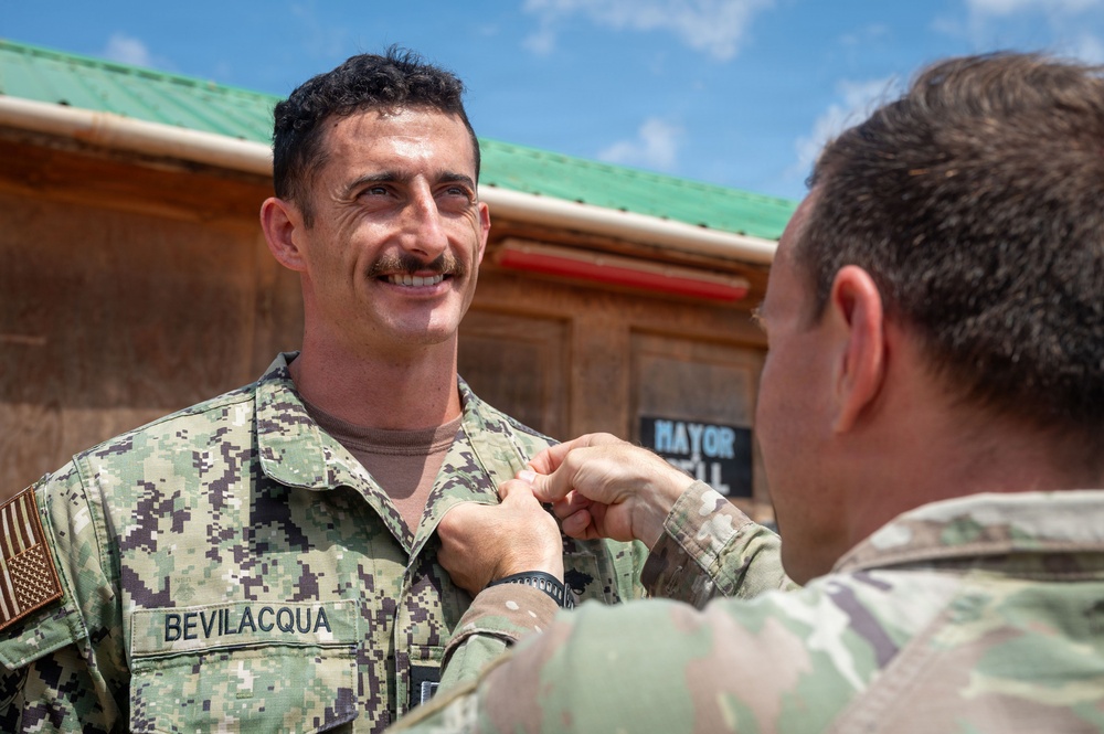 A military ceremony is taking place outdoors where one service member is pinning a medal or insignia onto another. The recipient, identified by his name tape as 'BEVILACQUA,' is wearing U.S. Navy Working Uniform (NWU Type III) with an American flag patch, and is smiling. The person performing the pinning is wearing U.S. Army OCP (Operational Camouflage Pattern) uniform.