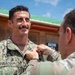 A military ceremony is taking place outdoors where one service member is pinning a medal or insignia onto another. The recipient, identified by his name tape as 'BEVILACQUA,' is wearing U.S. Navy Working Uniform (NWU Type III) with an American flag patch, and is smiling. The person performing the pinning is wearing U.S. Army OCP (Operational Camouflage Pattern) uniform.