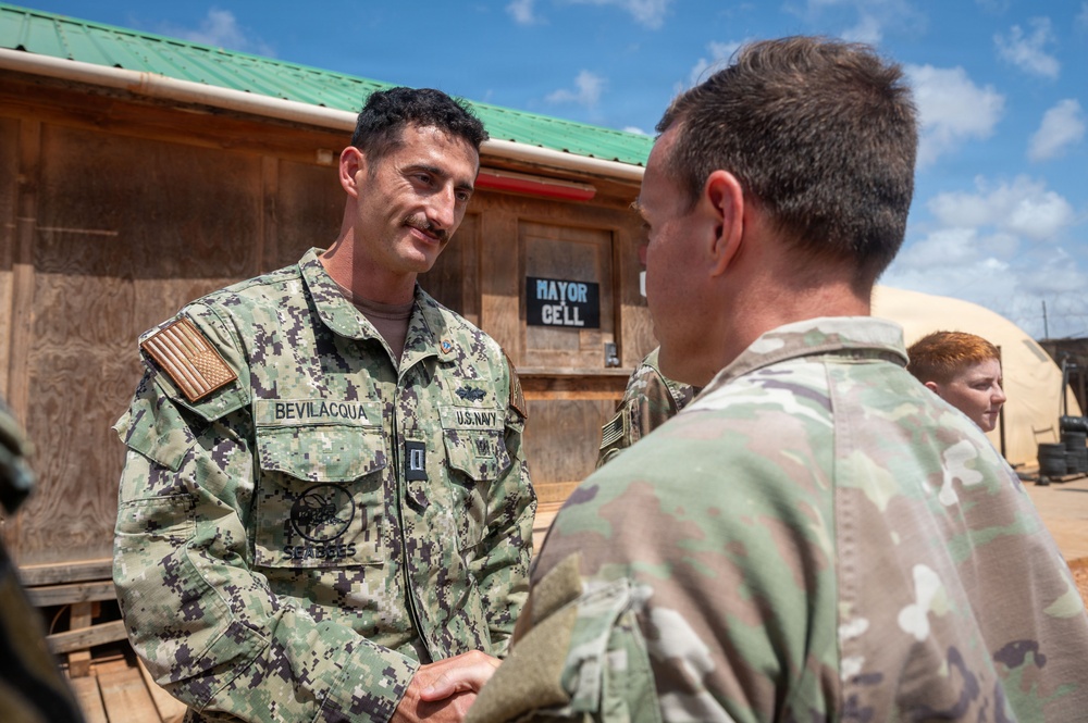 A U.S. Navy Seabee officer named Bevilacqua is shaking hands with a soldier wearing Army Combat Uniform (OCP/MultiCam pattern) at what appears to be a forward operating base or military installation. A sign reading 'MAYOR CELL' is visible on a wooden building in the background, indicating a base administrative area. A third service member with red hair is partially visible in the background.