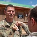 A military ceremony is taking place outdoors where one soldier is pinning something onto another soldier's uniform. The recipient, identified by his name tape as 'HOMES,' is a Sergeant First Class in the U.S. Army wearing OCP (Operational Camouflage Pattern) uniform, smiling during the ceremony. The setting appears to be a forward operating base or military installation, with a wooden building labeled 'MAYOR CELL' visible in the background.