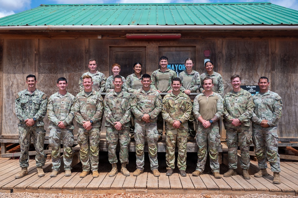 A group of approximately 15 military personnel posed for a formal group photo in front of a wooden building with a green metal roof. The group consists of two rows — a front row of approximately nine individuals and a back row of approximately six — all wearing military uniforms, predominantly Army OCP (Operational Camouflage Pattern) with one individual in Navy NWU (Naval Working Uniform). A sign on the building reads 'Welcome to Camp Tornado' or similar.