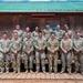 A group of approximately 15 military personnel posed for a formal group photo in front of a wooden building with a green metal roof. The group consists of two rows — a front row of approximately nine individuals and a back row of approximately six — all wearing military uniforms, predominantly Army OCP (Operational Camouflage Pattern) with one individual in Navy NWU (Naval Working Uniform). A sign on the building reads 'Welcome to Camp Tornado' or similar.