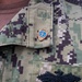 A close-up photograph of a military uniform collar area featuring a small shield-shaped pin with a blue background and gold border, displaying a figure and text. The uniform appears to be the US Navy Working Uniform (NWU Type I) in the distinctive digital woodland/urban camouflage pattern. A partial embroidered patch or badge is also visible on the chest pocket area.