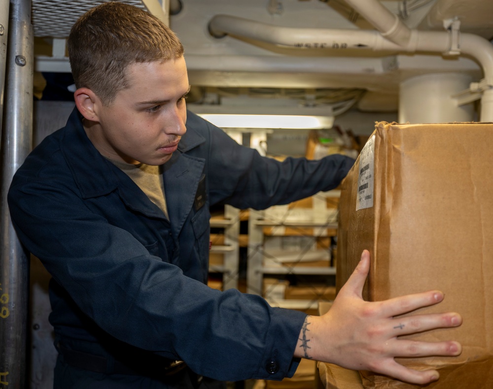 A young male service member in dark navy blue coveralls is handling a cardboard box in what appears to be a storage or supply area aboard a military vessel. He is focused on the box, which bears a shipping label. The background shows shelving units with additional packages and supplies, along with overhead pipes typical of a ship's interior.