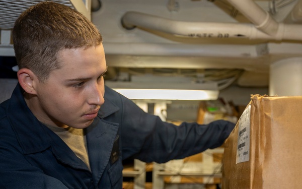 A young male service member in dark navy blue coveralls is handling a cardboard box in what appears to be a storage or supply area aboard a military vessel. He is focused on the box, which bears a shipping label. The background shows shelving units with additional packages and supplies, along with overhead pipes typical of a ship's interior.