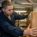 A young male service member in dark navy blue coveralls is handling a cardboard box in what appears to be a storage or supply area aboard a military vessel. He is focused on the box, which bears a shipping label. The background shows shelving units with additional packages and supplies, along with overhead pipes typical of a ship's interior.
