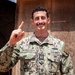 A U.S. Navy Seabees officer named Bevilacqua is photographed in NWU Type III camouflage uniform, holding up one finger and smiling. He appears to be outdoors in front of a plywood structure in what looks like a deployed or field environment. His rank insignia indicates he holds the rank of Lieutenant (O-3) in the U.S. Navy.
