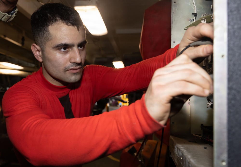 A young man wearing a red long-sleeve shirt is working on what appears to be mechanical or electrical equipment in an industrial or military setting. He is focused intently on manipulating components on a panel or machinery, using both hands. The environment appears to be an enclosed workspace with overhead fluorescent lighting.