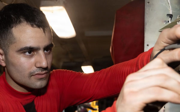 A young man wearing a red long-sleeve shirt is working on what appears to be mechanical or electrical equipment in an industrial or military setting. He is focused intently on manipulating components on a panel or machinery, using both hands. The environment appears to be an enclosed workspace with overhead fluorescent lighting.