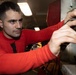 A young man wearing a red long-sleeve shirt is working on what appears to be mechanical or electrical equipment in an industrial or military setting. He is focused intently on manipulating components on a panel or machinery, using both hands. The environment appears to be an enclosed workspace with overhead fluorescent lighting.