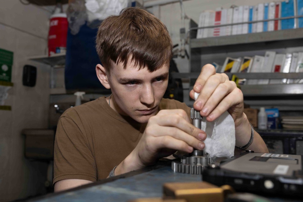 A young male technician is focused on assembling or disassembling a small mechanical component, likely a gear or bearing assembly, at a workbench. He is wearing a brown t-shirt and appears to be in a workshop or maintenance facility. The background shows shelving with binders, manuals, and various supplies.