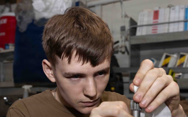 A young male technician is focused on assembling or disassembling a small mechanical component, likely a gear or bearing assembly, at a workbench. He is wearing a brown t-shirt and appears to be in a workshop or maintenance facility. The background shows shelving with binders, manuals, and various supplies.