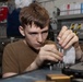 A young male technician is focused on assembling or disassembling a small mechanical component, likely a gear or bearing assembly, at a workbench. He is wearing a brown t-shirt and appears to be in a workshop or maintenance facility. The background shows shelving with binders, manuals, and various supplies.