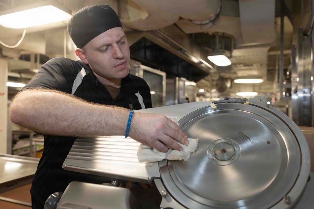 A male kitchen worker wearing a black cap and dark polo shirt is cleaning a commercial meat slicer with a white cloth in what appears to be a large institutional or commercial kitchen. He is focused on wiping down the circular blade guard of the stainless steel slicer. The background reveals an extensive industrial kitchen with stainless steel equipment, ductwork, and overhead lighting.
