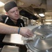 A male kitchen worker wearing a black cap and dark polo shirt is cleaning a commercial meat slicer with a white cloth in what appears to be a large institutional or commercial kitchen. He is focused on wiping down the circular blade guard of the stainless steel slicer. The background reveals an extensive industrial kitchen with stainless steel equipment, ductwork, and overhead lighting.