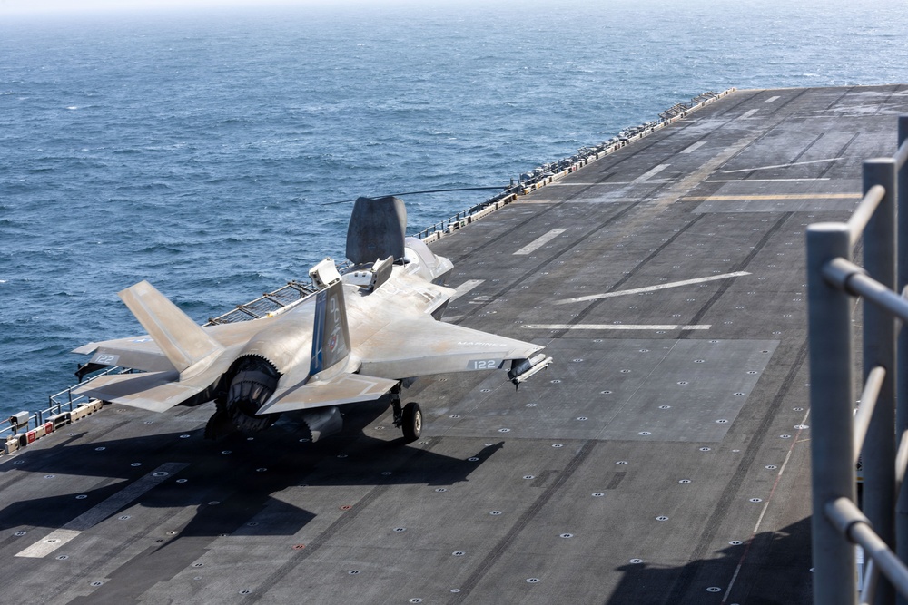 An F-35B Lightning II stealth fighter jet, marked with the number '122', is positioned on the flight deck of a naval vessel, likely an amphibious assault ship. The aircraft appears to be in a landing or taxiing configuration, with landing gear deployed and the lift fan door visible on the fuselage. The ship is underway at sea, with open ocean visible in the background.