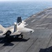 An F-35B Lightning II stealth fighter jet, marked with the number '122', is positioned on the flight deck of a naval vessel, likely an amphibious assault ship. The aircraft appears to be in a landing or taxiing configuration, with landing gear deployed and the lift fan door visible on the fuselage. The ship is underway at sea, with open ocean visible in the background.