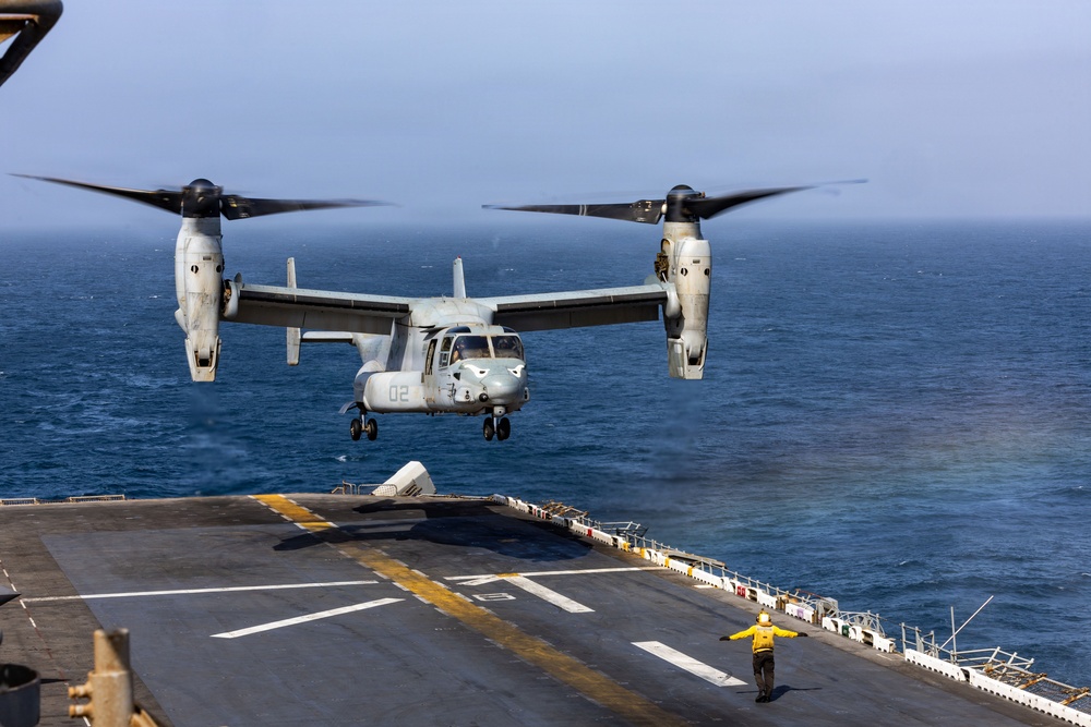 A U.S. Marine Corps MV-22 Osprey tiltrotor aircraft is captured mid-approach for landing on the flight deck of a naval amphibious assault ship at sea. The aircraft, marked with the number '02', has its rotors in vertical helicopter mode for the landing sequence. A flight deck handler in a yellow jersey is visible on the deck, providing visual guidance signals to the incoming aircraft.