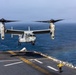 A U.S. Marine Corps MV-22 Osprey tiltrotor aircraft is captured mid-approach for landing on the flight deck of a naval amphibious assault ship at sea. The aircraft, marked with the number '02', has its rotors in vertical helicopter mode for the landing sequence. A flight deck handler in a yellow jersey is visible on the deck, providing visual guidance signals to the incoming aircraft.