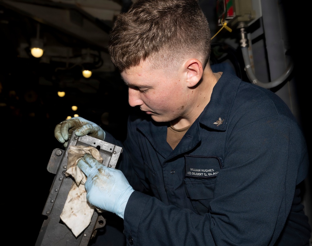 A U.S. Navy sailor identified as William Hughes, assigned to the USS Delbert D. Black, is performing maintenance work in what appears to be a ship's interior compartment. He is wearing blue nitrile gloves and a Navy working uniform (NWU) while cleaning or inspecting a metal mechanical component using a cloth rag. The background shows a dimly lit shipboard passageway or workspace with overhead lighting and piping.