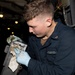 A U.S. Navy sailor identified as William Hughes, assigned to the USS Delbert D. Black, is performing maintenance work in what appears to be a ship's interior compartment. He is wearing blue nitrile gloves and a Navy working uniform (NWU) while cleaning or inspecting a metal mechanical component using a cloth rag. The background shows a dimly lit shipboard passageway or workspace with overhead lighting and piping.