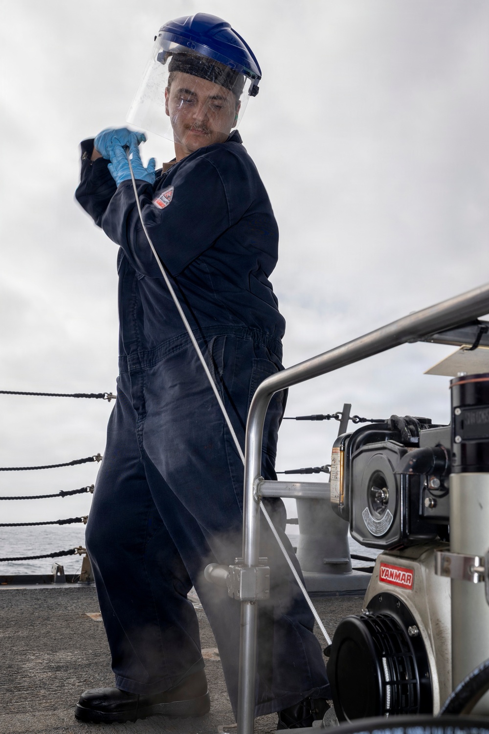 A worker in navy blue coveralls operates a Yanmar pressure washer on what appears to be the deck of a naval vessel. The individual is wearing personal protective equipment including a blue hard hat with a clear face shield and blue nitrile gloves. Steam or mist is visible near the machine, indicating active high-pressure washing operations.