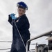 A worker in navy blue coveralls operates a Yanmar pressure washer on what appears to be the deck of a naval vessel. The individual is wearing personal protective equipment including a blue hard hat with a clear face shield and blue nitrile gloves. Steam or mist is visible near the machine, indicating active high-pressure washing operations.