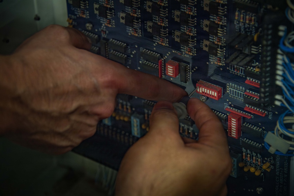 Two hands, appearing to belong to an older individual, are manipulating components on a blue printed circuit board (PCB). The person appears to be adjusting or configuring red DIP switches using what may be a small tool or fingernail. The board contains numerous electronic components including ICs, resistors, capacitors, and wiring harnesses visible on the right edge.