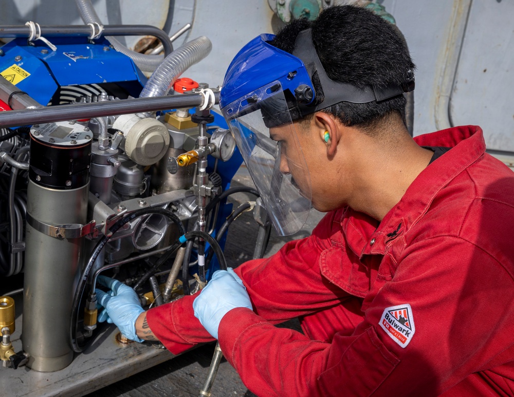 A technician wearing a red Bulwark FR (flame-resistant) coverall is performing maintenance or repair work on a complex mechanical assembly, likely an engine or compressor unit. The worker is equipped with a blue face shield, blue nitrile gloves, and ear protection, indicating adherence to proper personal protective equipment (PPE) protocols. The equipment being serviced appears to be an industrial or military-grade machine with multiple hoses, gauges, and metal components.