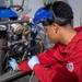 A technician wearing a red Bulwark FR (flame-resistant) coverall is performing maintenance or repair work on a complex mechanical assembly, likely an engine or compressor unit. The worker is equipped with a blue face shield, blue nitrile gloves, and ear protection, indicating adherence to proper personal protective equipment (PPE) protocols. The equipment being serviced appears to be an industrial or military-grade machine with multiple hoses, gauges, and metal components.