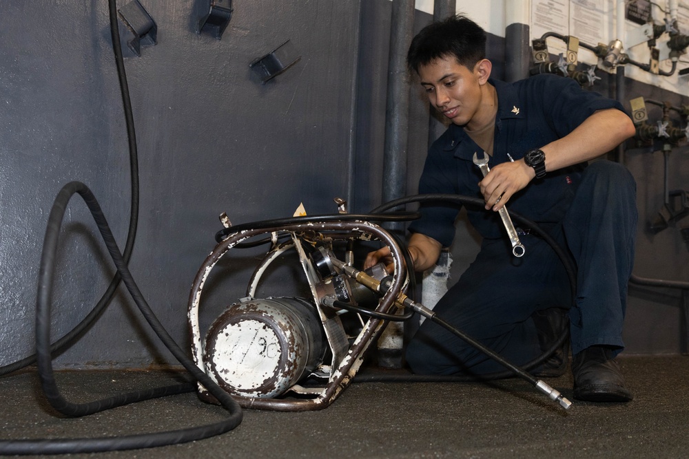 A young male in dark navy blue coveralls is crouched on the floor working on a piece of mechanical equipment with a wrench. The equipment appears to be a heavily worn hydraulic or pneumatic unit with hoses attached, located in what appears to be a naval vessel workspace. The individual is focused on the task, suggesting routine maintenance or repair work.