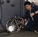 A young male in dark navy blue coveralls is crouched on the floor working on a piece of mechanical equipment with a wrench. The equipment appears to be a heavily worn hydraulic or pneumatic unit with hoses attached, located in what appears to be a naval vessel workspace. The individual is focused on the task, suggesting routine maintenance or repair work.