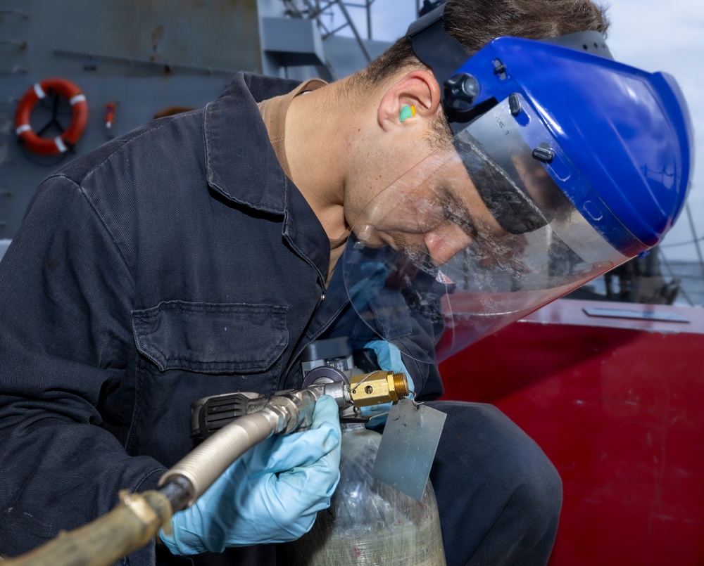 A worker wearing a blue face shield, blue nitrile gloves, and ear protection is performing a mechanical or pneumatic task on what appears to be a cylindrical metal component aboard a naval vessel. The worker is dressed in a dark navy coverall uniform and is using a pneumatic tool connected to an air hose. A life ring and ship superstructure are visible in the background, confirming a maritime/naval setting.