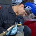 A worker wearing a blue face shield, blue nitrile gloves, and ear protection is performing a mechanical or pneumatic task on what appears to be a cylindrical metal component aboard a naval vessel. The worker is dressed in a dark navy coverall uniform and is using a pneumatic tool connected to an air hose. A life ring and ship superstructure are visible in the background, confirming a maritime/naval setting.