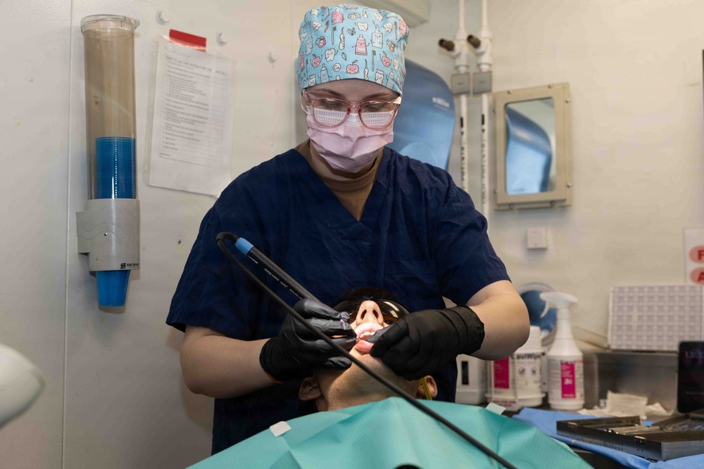 A dental professional in scrubs, surgical cap, face mask, glasses, and black gloves is performing a dental procedure on a reclined patient. The practitioner is using what appears to be a dental imaging or scanning device inside the patient's open mouth. The setting is a clinical dental operatory with various supplies and equipment visible in the background.