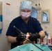 A dental professional in scrubs, surgical cap, face mask, glasses, and black gloves is performing a dental procedure on a reclined patient. The practitioner is using what appears to be a dental imaging or scanning device inside the patient's open mouth. The setting is a clinical dental operatory with various supplies and equipment visible in the background.