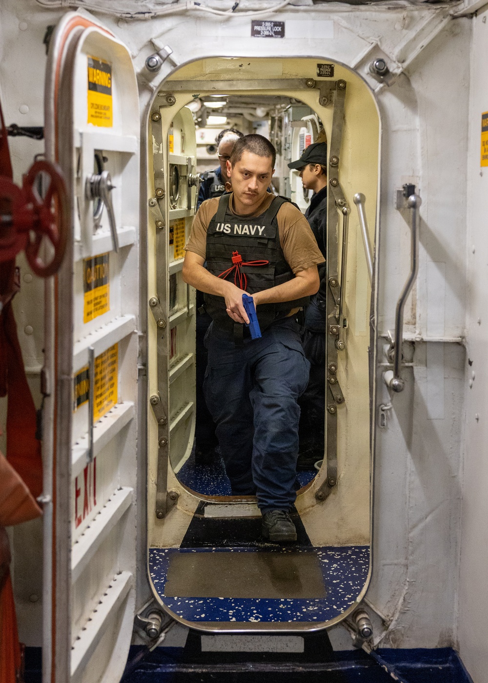 A US Navy sailor in a tactical vest is stepping through a watertight hatch/door aboard a naval vessel, holding a blue training pistol (simunition/dummy weapon). Additional personnel are visible in the passageway behind him, suggesting a training drill or security exercise. The setting is clearly the interior of a military ship, identifiable by the characteristic oval watertight door, warning labels, and naval interior fixtures.