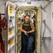 A US Navy sailor in a tactical vest is stepping through a watertight hatch/door aboard a naval vessel, holding a blue training pistol (simunition/dummy weapon). Additional personnel are visible in the passageway behind him, suggesting a training drill or security exercise. The setting is clearly the interior of a military ship, identifiable by the characteristic oval watertight door, warning labels, and naval interior fixtures.