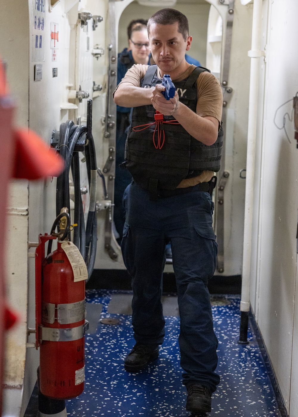 A U.S. Navy sailor in a tactical vest holds a blue training/simulation pistol in a two-handed shooting stance inside a narrow shipboard corridor. A second person in Navy uniform is visible in the background. The scene depicts a shipboard security or force protection training drill.