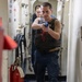 A U.S. Navy sailor in a tactical vest holds a blue training/simulation pistol in a two-handed shooting stance inside a narrow shipboard corridor. A second person in Navy uniform is visible in the background. The scene depicts a shipboard security or force protection training drill.