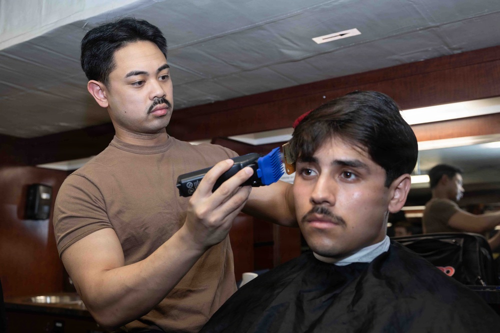 A barber is cutting the hair of a seated client in what appears to be a barbershop. The barber, wearing a brown t-shirt, is using electric clippers with a blue guard attachment on the client's hair. The client is draped in a black barber cape and appears to be looking forward calmly.